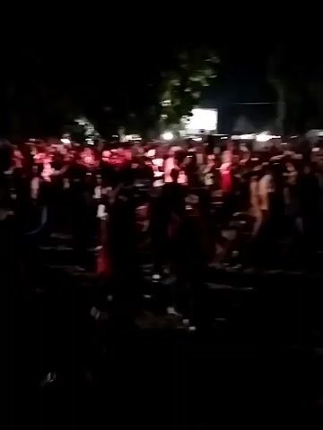 A crowd of people watch a performance on a stage. The stage is lit with bright lights, and there are speakers on either side. The crowd is dark, with only hints of light reflecting off people's clothing. Some people in the crowd are holding up their phones to record the performance. The performance appears to be a band or musical act. The overall atmosphere is one of excitement and engagement.
