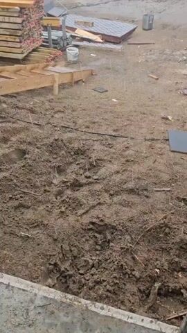 The video shows a construction site with stacks of lumber. In the background, solar panels are visible. The ground is muddy, and it appears to be raining. There are some construction materials scattered around, including what looks like roofing tiles. The camera is panning across the scene, giving a general overview of the site. No people are visible in the video.