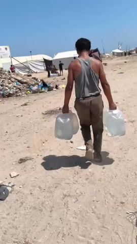 A person walks away from the camera on a dirt path, carrying two large plastic jugs of water. The person is wearing a grey tank top and brown pants. To the left of the path, there is a pile of trash and debris, including plastic bags, bottles, and some larger items. In the background, there are several white tents, some of which appear to be damaged. There are also some people visible near the tents. The sky is blue and the scene appears to be in a sunny, outdoor location, likely a refugee camp or similar setting.