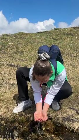 A young woman with a backpack crouches down beside a small stream on a grassy hillside. She is cupping her hands to drink from the water. The woman is wearing a white and green vest over a white long-sleeved shirt, black pants, and white sneakers. Her hair is pulled back in a ponytail with a scrunchie. She appears focused on drinking.

The scene is outdoors, with a hilly, grassy terrain under a bright blue sky dotted with fluffy white clouds. The vegetation appears dry and golden, suggesting a warm season, possibly summer or late spring. The sun is high in the sky, indicating it is likely midday. The location cues, such as the terrain and vegetation, suggest a mountainous or high-altitude environment. The overall mood seems peaceful and perhaps a bit adventurous, given the outdoor setting and activity. No visible text is present in the media. The estimated location is Raduil, Bulgaria.