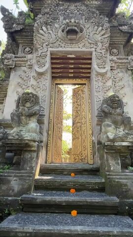 The video shows a set of ornate, golden doors opening to reveal a courtyard. The doors are elaborately carved with floral and peacock motifs. They are set in a stone structure flanked by sculpted figures. A few orange flowers are placed on the steps leading up to the doors. The courtyard beyond the doors features plants, paving stones, and another building with a thatched roof, visible in the distance. The perspective is from someone approaching the entrance, moving through the doors as they open.