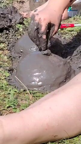 The video shows a close-up view of two mounds of wet, gray mud, likely formed by hands. A person's hands, covered in mud, are actively shaping the top of the right mound, pressing a small piece of dark material into it. Another person's legs and feet, also covered in mud, are visible in the foreground and background, suggesting they are also interacting with the mud. The setting appears to be outdoors, with some green grass and dry earth visible around the mud mounds. A red and white object, possibly a tool, is present in the background. The overall impression is of people playing or creating something with mud.