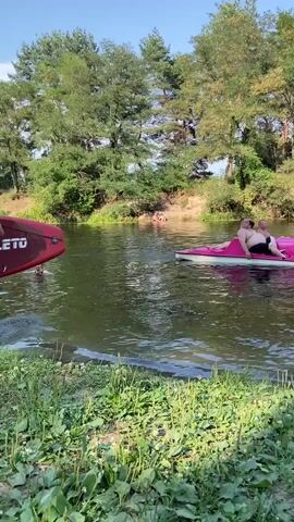 A man in blue shorts stands in shallow water next to a red paddleboard. He is partially submerged in the water. Behind him, a pink pedal boat with two people is on the water. In the background, there is a line of trees and a clear sky.