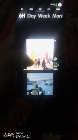 A hand scrolls through a series of short videos on a smartphone. The screen displays several small video previews arranged in a grid. The videos depict various scenes including a group of people walking in a crowd, a first-person view of someone skiing, a crowd of men in suits and traditional clothing, and a building in a body of water. The smartphone interface includes options like "6H," "Day," "Week," and "Mon," along with other icons and text. The phone is held against a dark, patterned surface. The phone model is identified as a Tecno Spark 9 Pro, displayed in the bottom left corner. The lighting is dim, focusing on the phone screen.