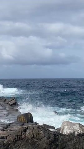 The video shows a close-up view of waves crashing against a rocky shore and a concrete breakwater under a cloudy sky. The water is a deep blue, and the waves are white with foam as they break against the rocks and concrete structures. The sky is filled with dark, heavy clouds, suggesting an overcast or stormy day. The foreground consists of dark, rough rocks, leading to the breakwater made of concrete blocks. The waves are the main action, creating a dynamic and powerful scene of nature. There are no people visible in the video. The sound of the crashing waves is audible.