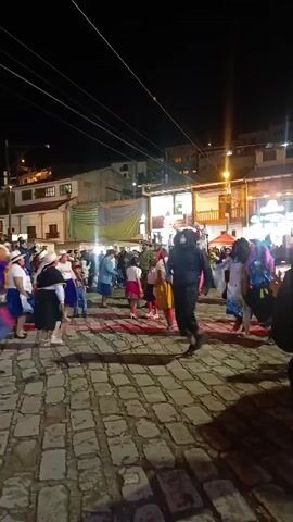 At night, on a cobblestone street, a group of people dressed in colorful costumes dance in a circle. There are buildings with lit windows in the background. The sky is dark, with streetlights and power lines visible. Some onlookers are present, observing the dancers and using their phones.