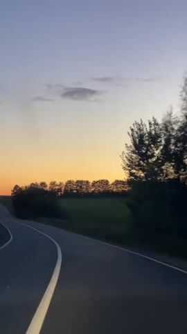 The video shows a drone shot from a low angle, moving forward along a winding paved road. The road has a white line down the center and curves to the left. On the right side of the road, there is a line of trees and vegetation. Beyond the trees, there is a field of green grass. The sky is a gradient of colors, starting with a deep blue at the top, transitioning to a pale yellow and orange near the horizon, indicating either sunrise or sunset. A few wispy clouds are visible in the sky. There are no people or other vehicles visible in the frame. The overall impression is of a serene, open landscape during twilight.