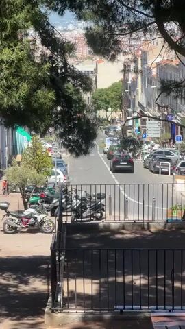 A slightly elevated view looking down a tree-lined street with buildings on either side. The street is busy with cars and motorbikes, and there are parked cars along the roadside. In the foreground, there is a black metal fence. The sun is shining and the sky is clear. The overall impression is a bustling urban scene on a sunny day.