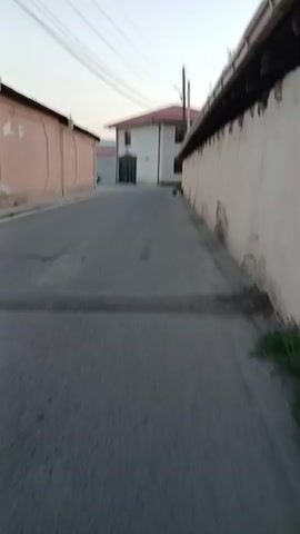 A person walks down an empty street with buildings on either side. There are power lines overhead and a car is parked in the distance. The ground is asphalt and there is some debris on the street.