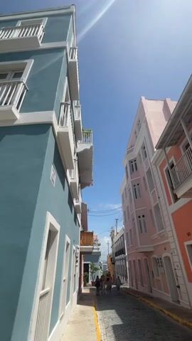 The video pans up from a cobblestone street lined with colorful buildings. A group of people walks down the street, appearing to be tourists. The buildings are painted in vibrant colors such as blue, pink, and orange, and feature balconies with white railings. The sky is a clear blue with some lens flare from the sun. The scene appears to be in Old San Juan, Puerto Rico.