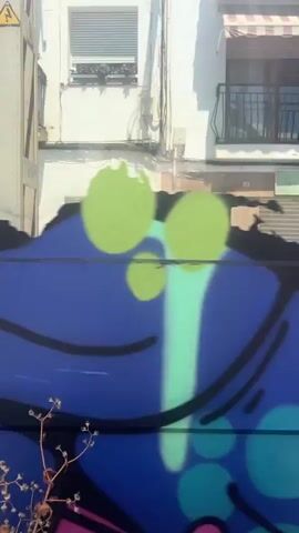The video opens with a reflection of a person taking a selfie in the window of a moving train, partially obscured by graffiti on the train's exterior. The person wears a hat and holds a smartphone. The train, which is orange and white with some graffiti, moves past the camera.

The scene then shifts to a view from the train window. In the immediate foreground are train tracks with gravel. Beyond the tracks, there's a narrow strip of sandy and rocky terrain, leading to the sea. The water is a clear turquoise-blue, stretching out to the horizon under a bright blue sky dotted with scattered white clouds. A solitary figure, dressed in red, stands near the water's edge, and an orange umbrella is visible on the sand a little further down. Overhead, power lines and a utility pole for the train are visible. The weather appears sunny and clear, indicating daytime. The sound of "woohoo" suggests a pleasant experience, likely enjoying the scenic coastal view from the train. This coastal setting aligns with the provided location context of Sant Pol de Mar, Spain.
