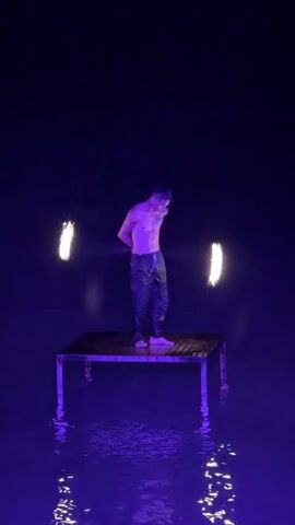 The media file captures a fire dancer performing on a platform surrounded by water. The setting appears to be at night, indicated by the dark sky and artificial lighting. The platform, possibly a table, is submerged in water, reflecting the purple and yellow lights from the fire. The performer, a shirtless young man wearing dark pants, is skillfully manipulating two fiery objects, likely poi or staffs, creating bright trails of light. His physique is lean and muscular, and his movements are fluid and deliberate. The overall mood is dramatic and mesmerizing, with the stark contrast between the darkness, the glowing fire, and the illuminated water creating an ethereal atmosphere. The location cues, Baan Chaweng, Thailand, suggest a tropical or coastal environment, possibly a beach or a lagoon, enhancing the exotic feel of the performance. There is no visible text in the media file.