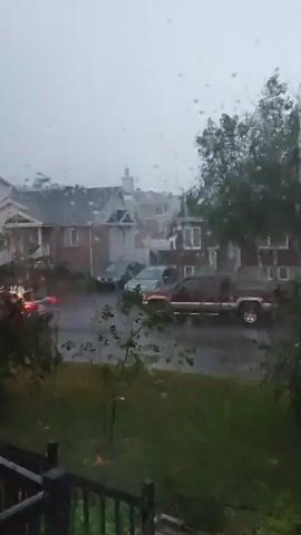 The video shows a residential street during a heavy rainstorm. Water droplets are visible on the window through which the video is filmed. Several houses with brick exteriors and white trim are visible. Parked in front of the houses are two cars and a red pickup truck. Trees and grass are also present in the scene. The overall impression is of a downpour with limited visibility due to the rain and overcast sky.