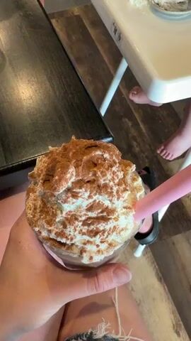 A first-person perspective video shows a hand holding a tall glass of a blended drink. The drink is topped with a generous amount of whipped cream and dusted with cinnamon. A pink straw is inserted into the drink. The background shows a casual dining setting, possibly a cafe or restaurant, with a high chair occupied by a small child in the distance. The child's feet are visible, and a small plate with some whipped cream and a pretzel is on the high chair tray. Other patrons are partially visible at a table to the left, with hints of furniture and decor suggesting an indoor environment. The lighting appears to be ambient, suggesting it could be daytime or evening indoors. The overall mood seems relaxed and casual, possibly a family outing.