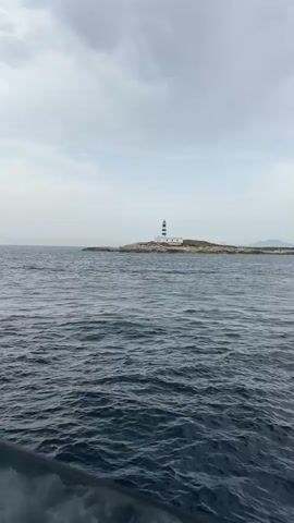 A first-person perspective video shows a person on a boat approaching a small island with a lighthouse. The water is dark blue with small waves. The sky is overcast. A hand enters the frame from the left, moving across the image. The hand is manicured with light-colored nail polish. The hand makes a waving motion at 00:01, points towards the lighthouse at 00:02, and then holds its palm open at 00:03 before disappearing from view. The lighthouse is black and white striped and stands on a rocky island with some vegetation.