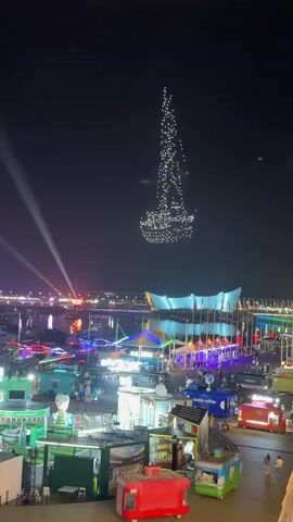 A drone show forms a silhouette of a sailboat in the night sky above a waterfront area filled with food stalls and entertainment. In the foreground, various colorful food kiosks are visible, some with people interacting around them. In the background, a large, modern building with a curved roof is illuminated, and lights from boats and other structures are scattered across the water. The drone formation is the central focus of the sky.