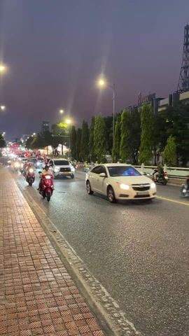 The video shows traffic in an urban area at night, likely in Vietnam based on the vehicles. The sky is a dark, dusky purple. Cars and a large number of motorbikes are driving on a road that is wet from rain. On the right side of the road there are tall, thin trees. On the left there is a paved sidewalk. Overhead street lights illuminate the road. The traffic seems heavy but moving.