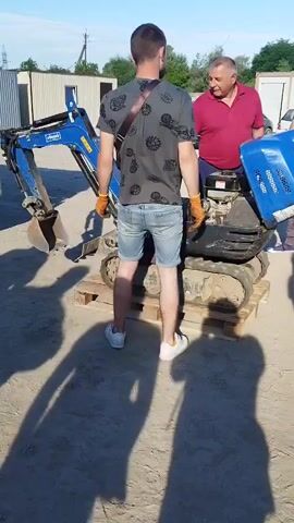A man wearing a dark t-shirt with a grey circular pattern, blue denim shorts, orange gloves, and white sneakers stands facing a small blue excavator. He has a dark bag strap across his chest. Another man in a red polo shirt stands to his right, also wearing an orange glove on his left hand. The excavator is on wooden pallets on a dirt or gravel surface. In the background, there are white buildings or shipping containers, some trees, and a truck. The sun is casting long shadows. The video is a short loop showing the same scene.