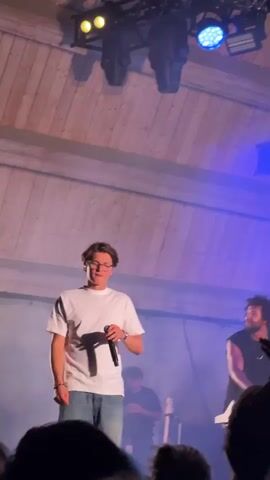 The video opens with a man in a white t-shirt and jeans singing into a microphone on a stage. He is wearing glasses and has light brown hair. Behind him, another man with dark curly hair is seated at a keyboard. The stage is lit with bright lights, and there is smoke or haze in the air. The background is a wooden ceiling structure. The camera then pans out to show a large crowd of people gathered in front of the stage, many holding up their phones to record. The scene transitions to a wider shot of the crowd at night, with trees and lights visible in the background. Some trees are illuminated with colored lights, including pink and green. The sky is dark, and the overall atmosphere appears to be that of an outdoor concert or festival.