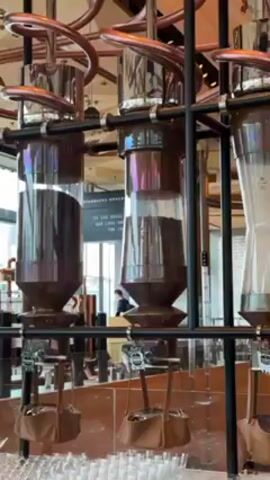 Here is a detailed description of the video:
The video shows a close-up view of a coffee roasting and brewing apparatus at a Starbucks Reserve Roastery. The equipment is comprised of large, cylindrical glass containers filled with coffee beans at various stages of the roasting or brewing process. These containers are arranged in a row and are part of a larger, complex system of pipes and supports made of metal, possibly copper or stainless steel. Underneath each container, there are small leather bags, presumably to collect the coffee after it has been processed. Behind the equipment, a blurred background reveals a cafe-like setting with tables, chairs, and other patrons. A sign with "STARBUCKS RESERVE" is partially visible in the background. The lighting is bright, illuminating the scene and highlighting the metallic and glass surfaces of the coffee equipment.