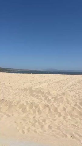 The video shows a sandy beach under a clear blue sky. The foreground is dominated by textured sand dunes. In the middle ground, a person is walking across the sand. In the background, the ocean is visible, with a distant shoreline and mountains. A red car is partially visible in the lower right corner of the frame. The person in the middle ground appears to be walking from left to right.