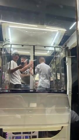 Three men are in a cabin of a Ferris wheel. The cabin is white with transparent windows and a lit roof.  The man closest to the camera is wearing a white hat with a black brim, sunglasses, and a white t-shirt. He is holding a phone and filming. The second man is wearing a black t-shirt and jeans. The third man is wearing a white collared shirt and jeans.  The Ferris wheel cabins pass by illuminated in red, blue, and green. The men are talking and interacting. The background is dark, suggesting it is nighttime.