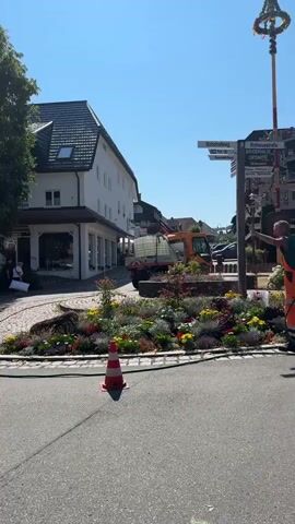 A man in an orange jumpsuit and green vest waters a flower bed with a hose. He stands on a paved street, facing away from the camera and towards the flower bed. In the background, there is a building, a signpost, and a small orange tractor. The sky is blue and clear. The man is using a green hose to water the plants. There are orange traffic cones on the street. The flower bed contains a variety of colorful flowers and plants.