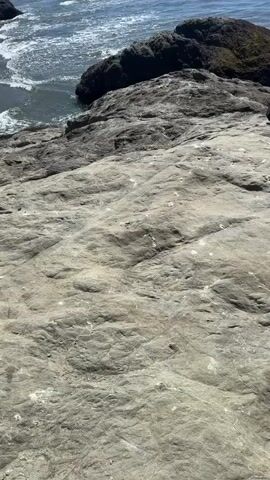 This is a POV shot of a person sitting on a large, light-colored rock formation at the edge of the ocean. The person is wearing dark clothing and is facing away from the camera, looking out at the water. The ocean is a deep blue with white waves crashing against the shore and around large, dark rock outcrops in the distance. The sky is a clear, bright blue. The camera is tilted upwards, showing the expansive ocean and sky.