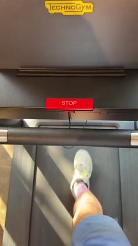 The video shows a first-person perspective of someone walking on a treadmill. The treadmill is a Technogym brand. The camera is pointed downwards at the treadmill belt and the person's legs and feet. The person is wearing blue shorts and white sneakers with colorful accents. The person is also wearing white socks with purple stripes. The treadmill has a red emergency stop button. The screen of the treadmill is visible at the top of the frame, showing speed and other workout metrics. The lighting suggests an indoor gym environment.