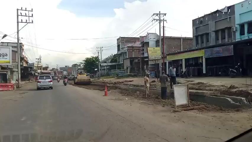 The video shows a street scene under construction. In the foreground, the edge of a vehicle is visible. A paved road runs through the scene, with ongoing construction work on the right side. A yellow steamroller is actively compacting the ground, with a person operating it. Several people are standing around the construction site, some appearing to be workers. Various buildings line the street, including shops with awnings and upper floors. Traffic is present, with a white car and a red motorcycle visible on the road. Construction materials and debris are scattered around the work area. Traffic cones are also present to mark off the construction zone. The overall environment suggests an active road repair or upgrade project in a commercial or residential area.