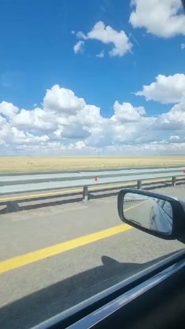 The media file shows a view from inside a moving car on a highway, looking out the passenger window and side mirror. The scene is a vast, flat landscape under a bright blue sky dotted with fluffy white clouds. The road has a yellow line painted on the right side, indicating a lane divider. A metal guardrail runs alongside the highway, separating it from a field of dry, yellowish grass that stretches to the horizon. In the distance, a few utility poles or small structures are visible. The side mirror reflects the road behind the car, showing another vehicle and the continuation of the highway. The time of day appears to be daytime, with clear, sunny weather. The overall impression is one of travel through an open, possibly rural or semi-arid region. No people are visible, and there is no visible text.