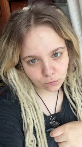 This is a close-up selfie video of a young woman with blonde dreadlocks. She is wearing a black t-shirt and a silver necklace with a green pendant. Her fingernails are long and painted a shimmery blue-green color. She has light skin with freckles and is not wearing makeup.

The woman is looking down at her finger, which is touching her lips, with a thoughtful expression. She then looks up at the camera, winks, and smiles. Her expression changes from pensive to playful and friendly. She then lightly touches her hair or necklace.

The background is softly blurred, but suggests an indoor setting. There is a hint of a white curtain on the right side of the frame, and a darker area on the left which might be furniture or a wall. The lighting appears natural, possibly from a window, suggesting it is daytime.

The video is very short, lasting only a few seconds, and appears to be a casual personal recording. There is no visible text in the video. The subject's demeanor is relaxed and engaging. The location context of Vostochny District, Russia is not visually discernible from the video content.