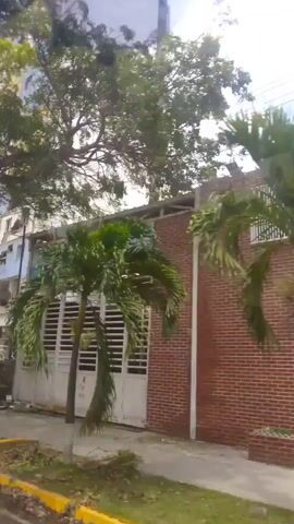 The video shows a multi-story building with significant structural damage. Sections of the facade have collapsed, revealing exposed interior spaces and debris scattered on the ground. The building's exterior is painted in alternating bands of blue, yellow, and white. A small blue van is parked in front of the building. There are trees and foliage around the building, and a pole with wires is visible. A person is walking away from the camera on a paved surface. The scene suggests a recent event such as an explosion or structural failure.