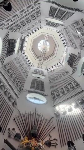 The camera looks up an octagonal shaped tower decorated with medieval weapons mounted on its walls. The weapons include spears, swords, shields, and helmets. There is a coat of arms at the bottom of the tower.  A chandelier hangs from the center of the octagonal ceiling at the top.  There are also several small round windows on the walls of the tower.