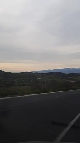 The camera is moving along a paved road, the road filling the right side of the view.  To the left of the road is a landscape of green hills, the terrain gradually sloping upward to form mountains in the distance. The mountaintops are faintly visible in the soft light of the sky. The sky itself occupies the left half of the image, featuring a mixture of grey clouds and clear patches. The camera is angled vertically. There is audible music in the background.