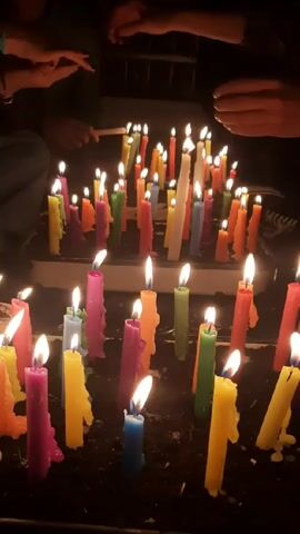 The video shows a close-up of a group of people lighting a collection of colorful candles in a dark environment. There are numerous candles of varying colors, including yellow, pink, green, orange, and blue, all planted in dark soil. Each candle has a lit flame. Several hands, some with painted nails, are visible around the arrangement, using a long match to light the candles. The scene appears to be focused on the act of lighting the candles, emphasizing the warm glow and color of the flames against the dark backdrop. There's some dripping wax visible on the sides of the candles, indicating that they have been burning for a while.