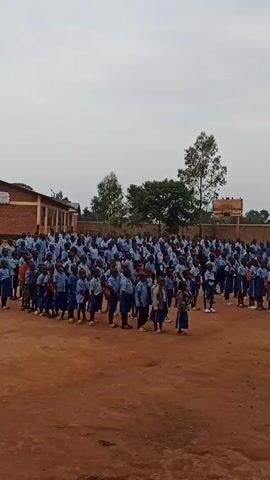 In a medium shot, a large group of school children dressed in matching light blue uniforms are standing in lines on a brown dirt field. They appear to be assembled in a schoolyard, with a single-story brick school building visible on the left and a brick wall in the background on the right. Several trees are scattered throughout the background. 

A man in a white shirt and dark pants stands to the right of the group, speaking to the children. The sky is overcast and gray. The camera is static and does not move throughout the short video. The overall impression is of a school assembly or gathering.