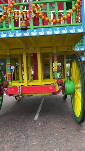 The video shows a colorful, wooden float decorated with yellow, blue, red, and green. The float has two large wheels, one prominently in the foreground, with yellow spokes and green rims. The sides of the float are adorned with a repeating pattern of white swans. Garlands of orange and yellow flowers hang from the top of the float. People are visible on the float and walking alongside it. Some individuals on the float appear to be in traditional attire. Several people walking beside the float are wearing high-visibility vests, suggesting they might be event organizers or participants in a procession. The ground is asphalt, marked with a white line. The background shows buildings and a cloudy sky. The camera is positioned low, focusing on the wheels and lower structure of the float as it moves.