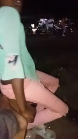 Two individuals are visible in a dimly lit outdoor setting. A person with dark skin, wearing a light blue-green patterned top and pink pants, is initially seen from the chest down, sitting or lying with their legs bent. Their right hand rests on their thigh. A second person's leg, clothed in grey pants, is partially visible next to the first individual's leg. The person in pink pants then sits upright. The camera subsequently pans down, focusing on a bare foot, presumably belonging to the second individual, stepping onto a wooden plank on the ground. The background is dark with some blurred lights in the initial frames.
