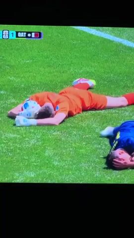 This media file shows a soccer match in progress. In the foreground, two players are lying on the grass, seemingly injured or exhausted. The player in the orange jersey is on his back with his head resting on a soccer ball. Another player in a blue jersey is lying on his side, holding his head. A third player in a white jersey with the number 11 on the back is standing over the player in the orange jersey, appearing to check on him. The scene is set on a bright, sunny day with a well-maintained green grass field. The scoreboard at the top left corner of the frame indicates that the score is 1-0, with "QAT" being one of the teams. The overall impression is one of a dramatic moment in a competitive soccer game, possibly following a collision or a moment of intense effort.