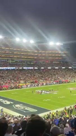 The video shows a football game in progress at a large, crowded stadium at night. The bright stadium lights illuminate the field, where players in white uniforms are lined up. The stands are full of spectators. "SEATTLE" is visible on the end zone.
