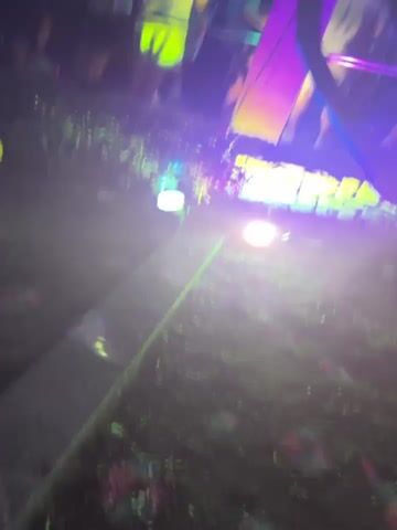 The video shows a nighttime outdoor gathering with people standing and moving. The scene is illuminated by purple and green lights, with some lights also appearing white. Trees with branches and leaves are visible in the foreground and mid-ground, with some branches obscuring parts of the scene. The ground appears to be made of stone. In the background, there are more people and structures that suggest a bar or club environment. The atmosphere appears to be festive, with the lighting creating a vibrant and dynamic visual effect.