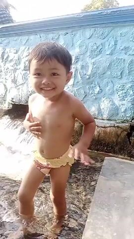 A young child, likely a toddler, stands in shallow water with a bright blue, textured wall behind them. The child is wearing yellow swim shorts with cartoon characters. They are looking to the left and smiling. In the background, there are several other people, some sitting on the wall, and others standing further away. The scene appears to be outdoors, with sunlight suggesting it is daytime. The presence of water and the casual attire of the people indicate a recreational setting, possibly a pool or natural water feature. The overall atmosphere seems relaxed and joyful, given the child's expression. There is no visible text.