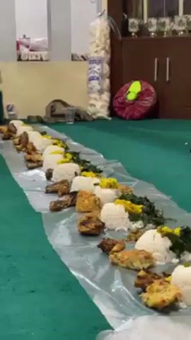 Here is a detailed description of the video:

The video shows a long, low-angle shot of a meal spread out on a plastic runner on a green carpet. The meal appears to consist of individual servings of white rice, fried chicken, vegetables, and what looks like fried tempeh or tofu. The servings are arranged in a linear fashion down the length of the plastic runner.

In the background, there's a cabinet with what appear to be decorative items on top of it. To the left of the cabinet, there are some bags and other items on a shelf, and in the background, a stack of packaged crackers.

A man wearing a white cap and glasses appears in the frame to the left of the meal. He raises two fingers in a peace sign, smiling at the camera. Another person can be seen in the lower right corner, just an arm and some clothing.

The lighting appears to be indoor, and the room looks to be a common area, possibly in a home or community center.