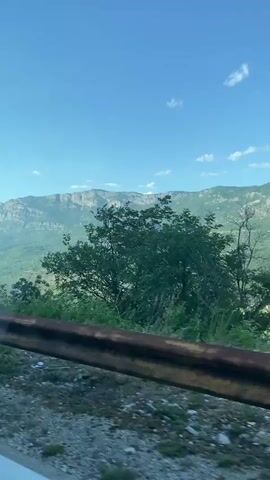 This is a video taken from a moving vehicle, likely a car, on a winding road in a mountainous region. The video begins with a shot of a triangular warning sign indicating falling rocks, followed by a view of lush green mountains under a clear blue sky. As the vehicle moves, the scenery changes, showing trees and the road ahead. The road appears to be paved with some gravelly sections and has a white line marking its edge. A rusty guardrail runs alongside the road for a portion of the video. The overall impression is of a scenic drive through a natural landscape.