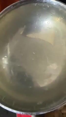 A metal bowl is shown from a top-down perspective. It is empty and reflects the surrounding environment. The bowl is then tilted, and water is poured from it into another metal bowl filled with dry kibble. The kibble pieces are small and round. As the water is poured, it saturates the kibble, causing some of it to clump together and darken in color. The video ends with a close-up of the kibble, now wet and glistening.