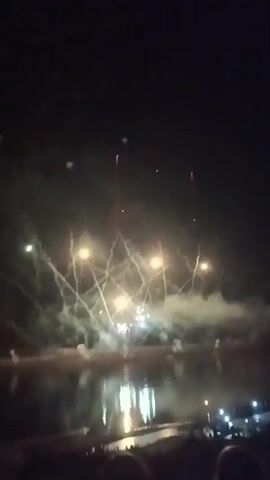 The video shows a nighttime fireworks display over a body of water. The fireworks are exploding in various colors, including red, pink, and blue. Some of the fireworks are creating large, spherical bursts, while others are producing trails of sparks. Smoke from the fireworks is visible in the air, and the lights of a distant city are reflected in the water. There are some silhouetted figures visible in the foreground, suggesting that people are watching the display. The overall atmosphere is festive and celebratory.