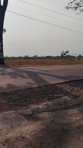 The video shows a road scene. On the left side, there are power lines and a tree branch. The road itself is paved, with a dirt or gravel area to the right. In the distance, there are fields and some buildings. A car drives by from left to right, followed by a motorcycle with a rider. The overall lighting suggests it is daytime, possibly morning or late afternoon due to the long shadows. The camera angle is tilted, giving a vertical perspective of the scene.