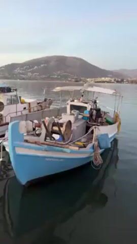 The video shows two fishing boats docked in a harbor. The first boat is white with a blue tarp roof and yellow netting filling its deck. The second boat is smaller, with a light blue hull and a cluttered deck. A hill and buildings are visible in the background across the water. The sky is overcast.