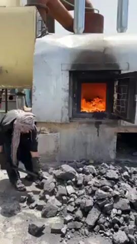 In an outdoor industrial setting in Kandahar, Afghanistan, a man is engaged in manual labor, fueling a large furnace. The scene is brightly lit by daylight, suggesting it is daytime.

The man, dressed in dark, loose-fitting clothing, a head covering, and protective gloves, stands beside a large pile of black coal chunks. He uses a long-handled shovel to scoop the coal. To his right, a sizable industrial furnace or boiler dominates the frame. Its metallic, possibly insulated, exterior has an open door revealing a roaring, intense fire burning brightly inside, indicating high temperatures. Various pipes and other metallic components are visible on and around the furnace, suggesting its industrial function.

The primary activity is the man repeatedly scooping coal from the pile on the ground and forcefully throwing it into the open, fiery mouth of the furnace. This action implies he is feeding the furnace to maintain its heat and operation. The ground appears to be a mix of dirt and concrete, covered with scattered coal dust and small fragments. The man's posture and movements suggest exertion and a rhythmic, practiced motion required for this demanding task. No visible text or specific brandings are present on the equipment.